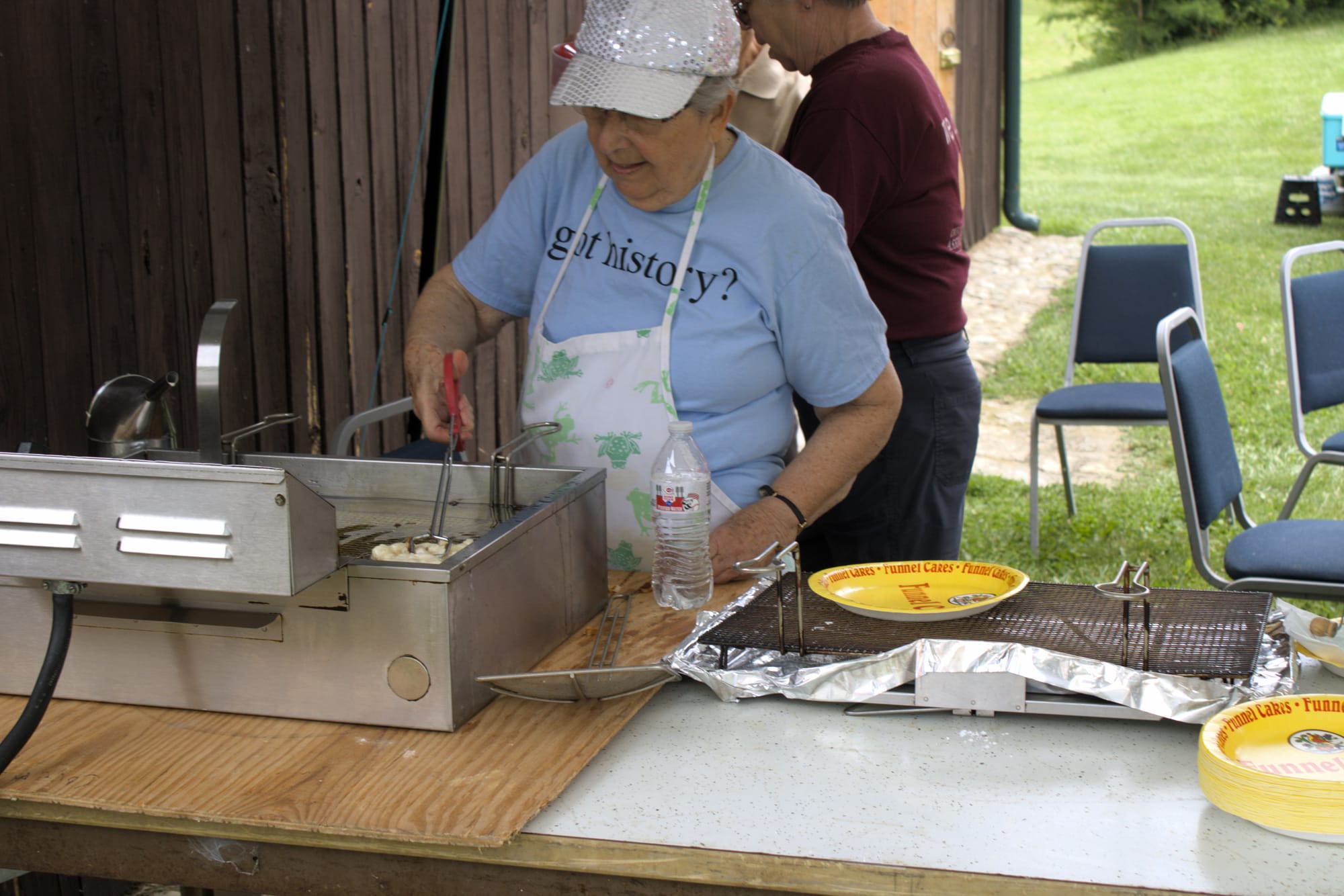 Woman in blue fries funnel cakes using a fryer on a wooden table.