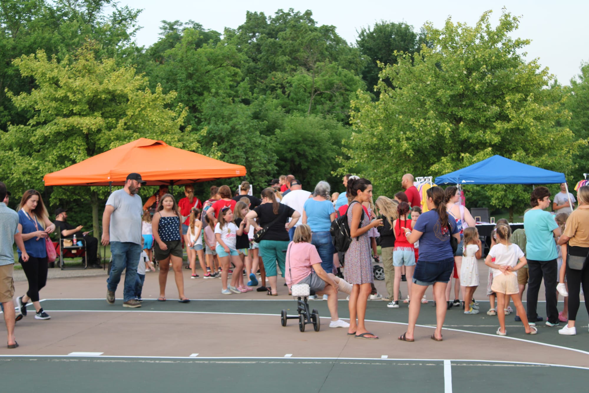 Dozens of people wait in line and walk around between booths set up on a basketball court outside