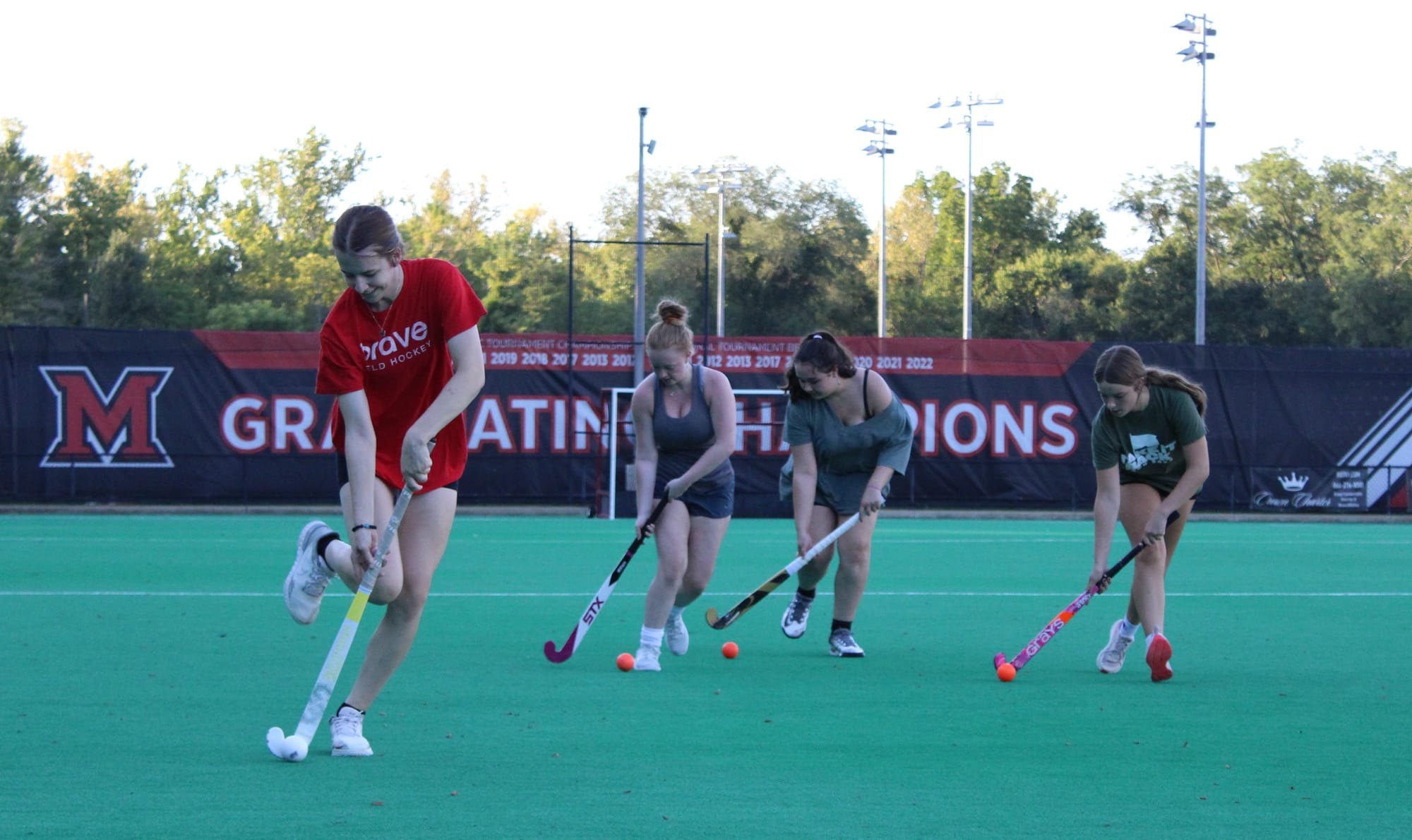 Four girls with field hockey sticks push balls in front of them on a field