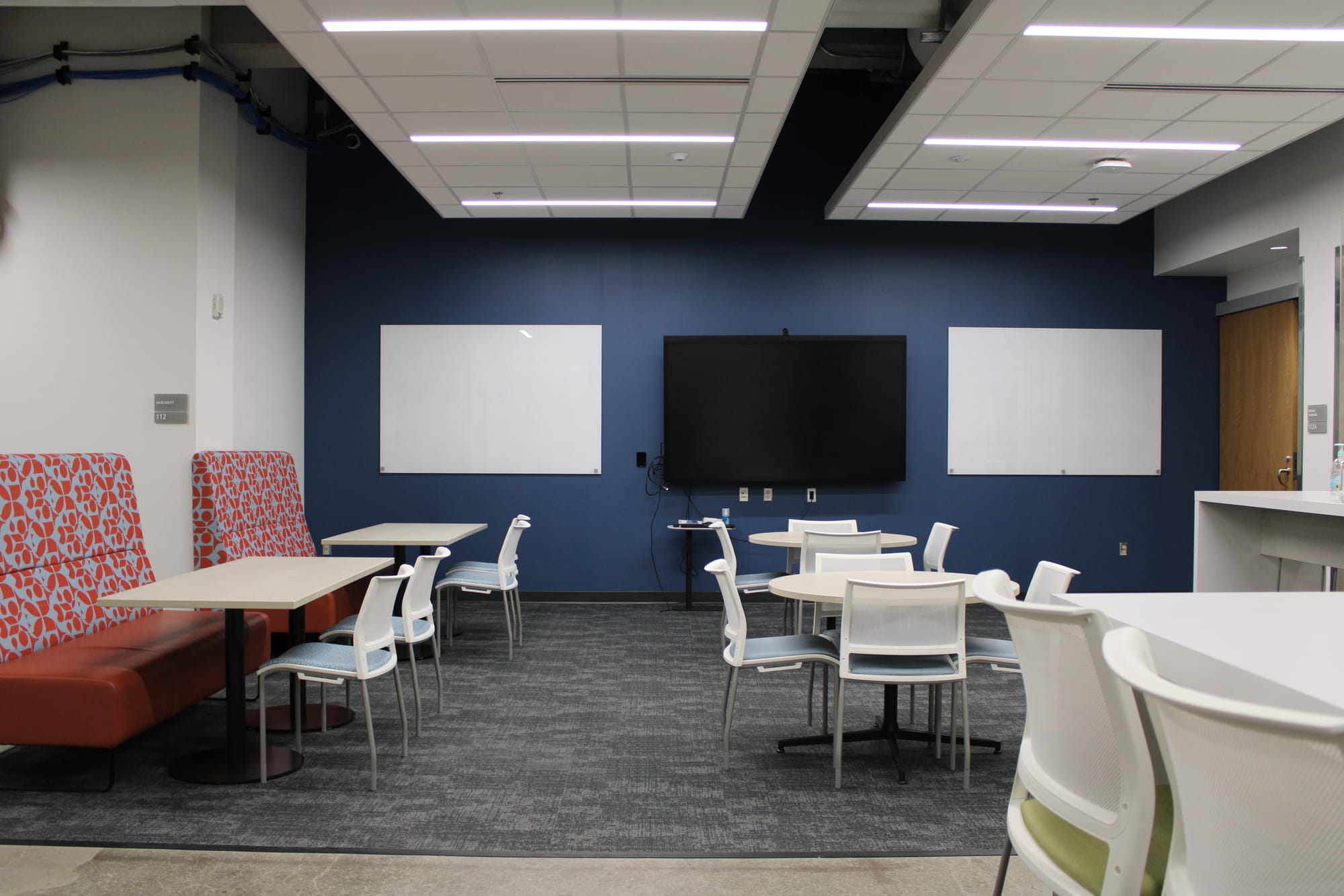 A room with several tables and chairs, two whiteboards and a television