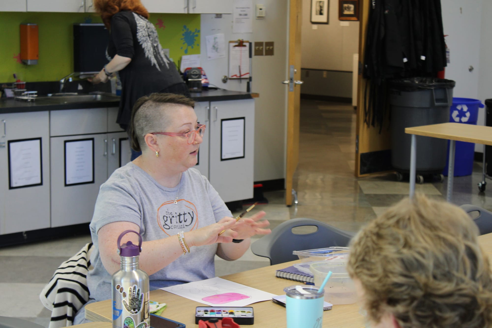 Janice Hudson sits at a table and speaks, paint brush in hand