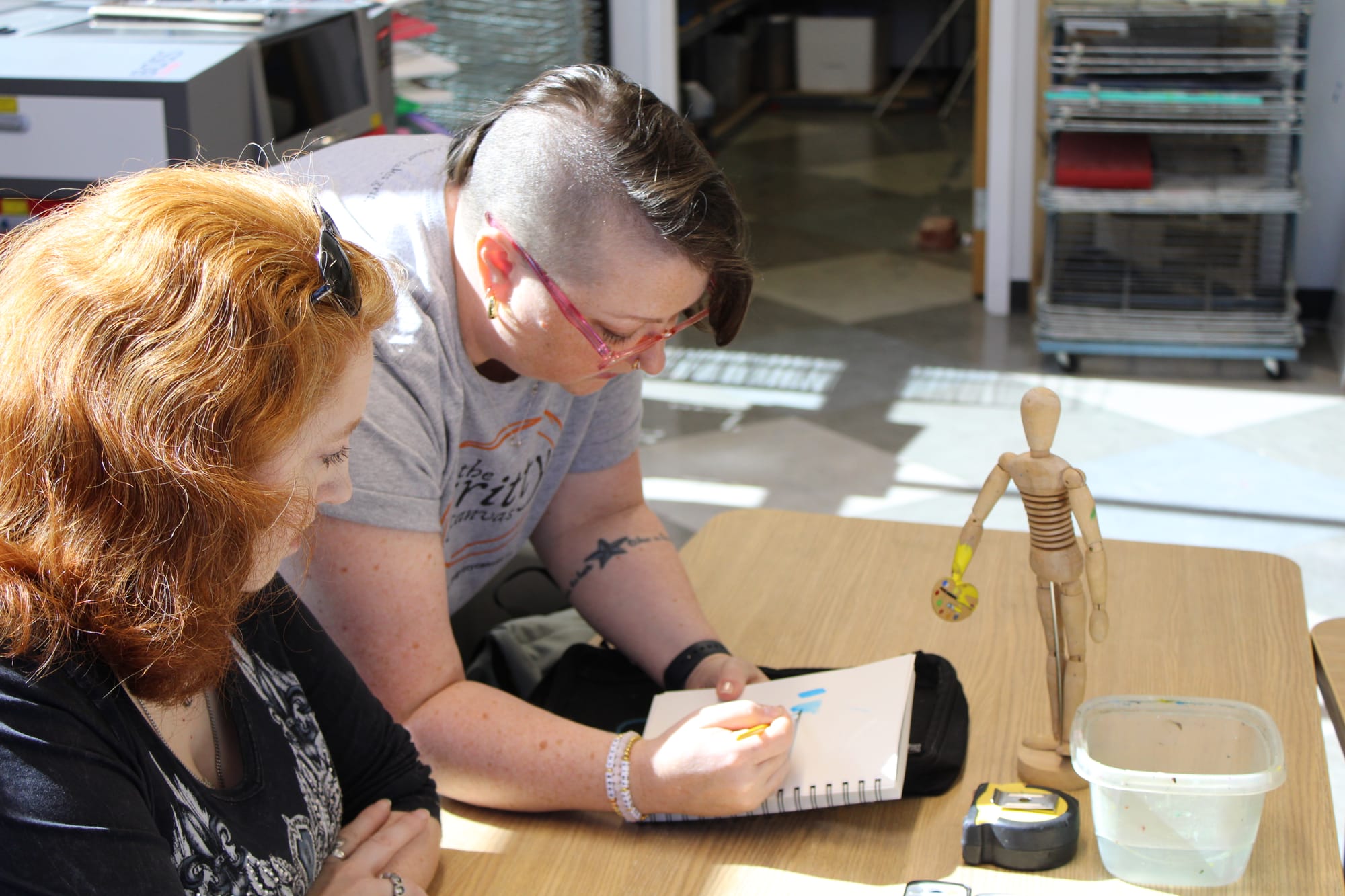 Janice Hudson stands to the right of Jennifer Sergent and paints in a notebook