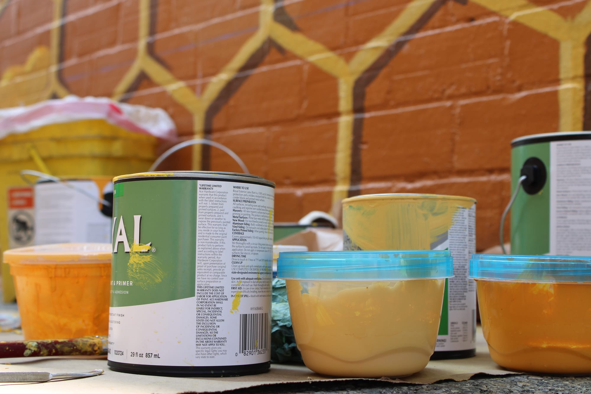 A variety of paint cans and tupperware with yellow and orange paint sit on the ground