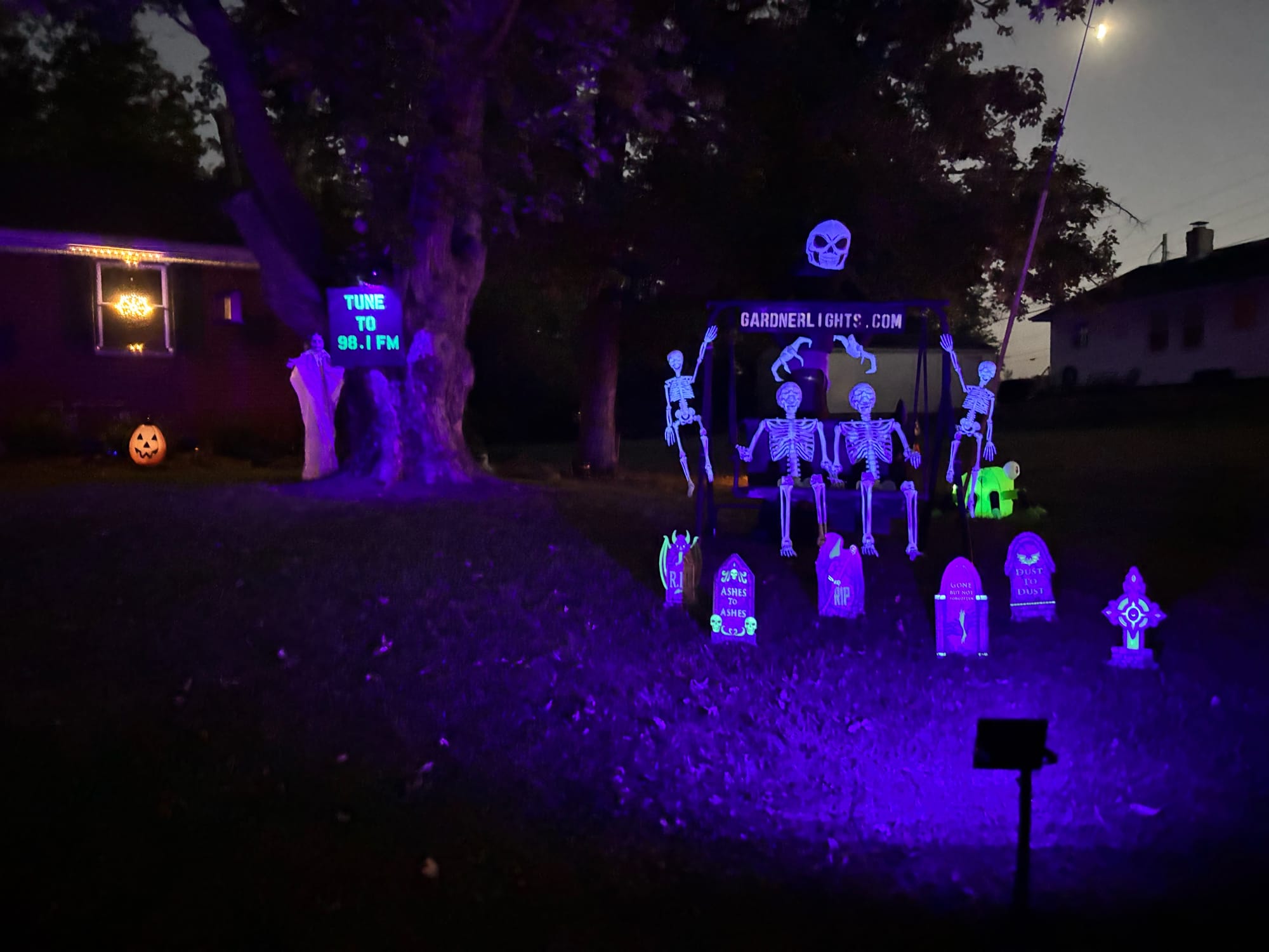 A view of a display reading gardnerlights.com with skeletons and tombstones in front of a tree reading "tune to 98.1 FM"