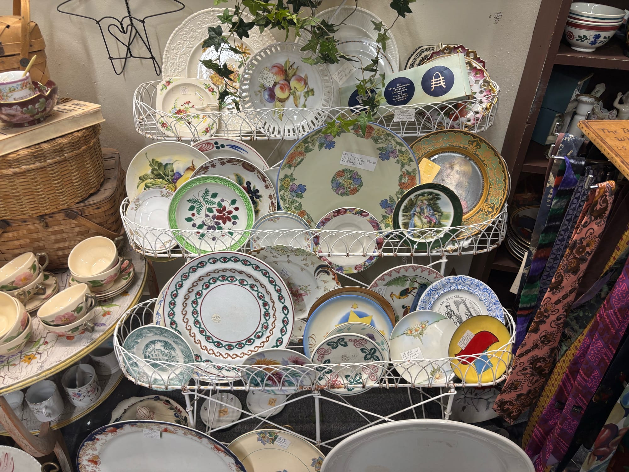 Antique plates on display on a three-tier shelf, next to a rack of ties on the right and tea cups on the left