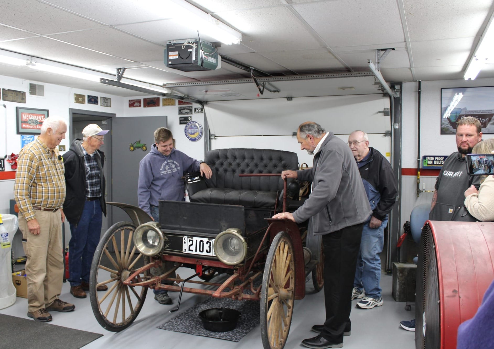 Several people stand around a 1911 Sears Motor Buggy, a car with large wooden wheels and no roof