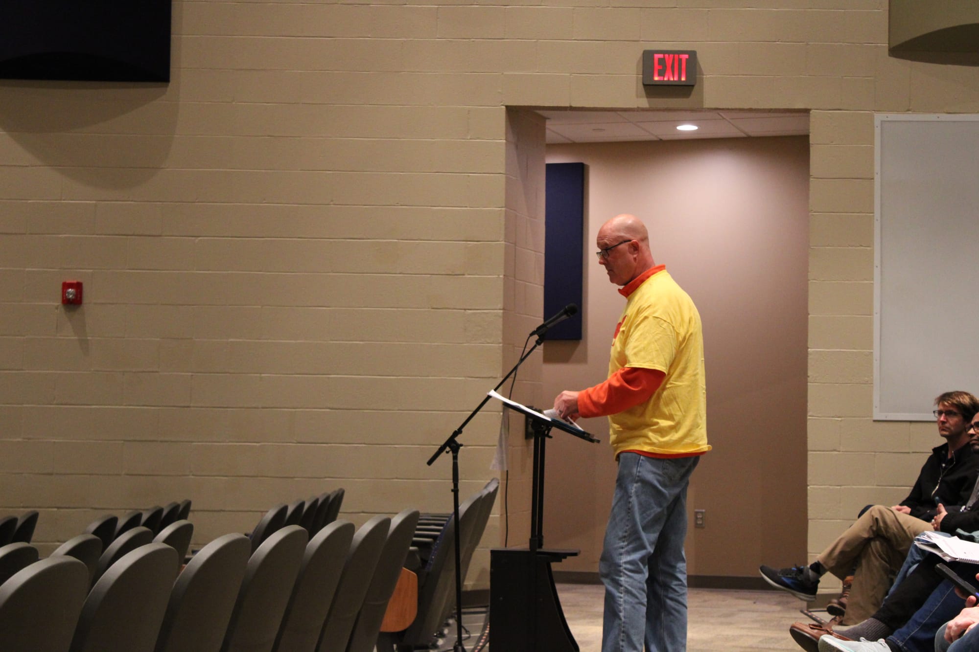 Art Sauerwein stands at a microphone in an auditorium wearing a yellow shirt