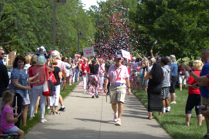 A man walks down a sidewalk with spectators on both sides. Behind him, more people are walking under confetti and holding si