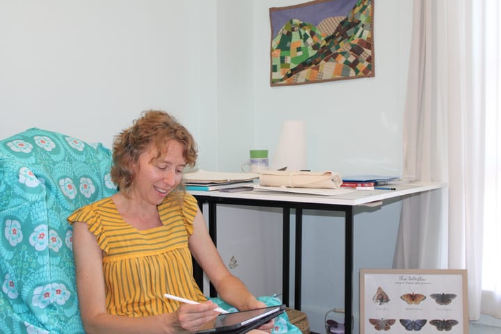 Amy Bartel sits in her studio. Her work table is filled with papers behind her, and art hangs on the walls