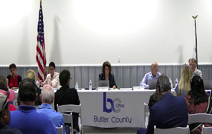 Five commissioners, three women and two men, sit at long white table in front of white and grey wall