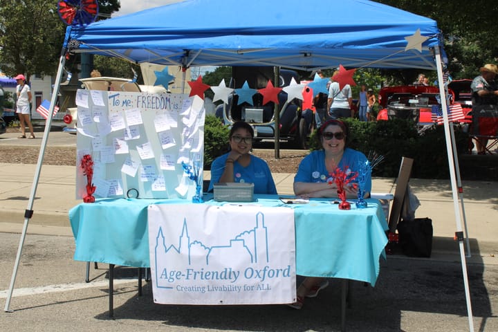 Jennifer-Heston Mullins and a second volunteer sit under a canopy with a table labeled "Age-Friendly Oxford"