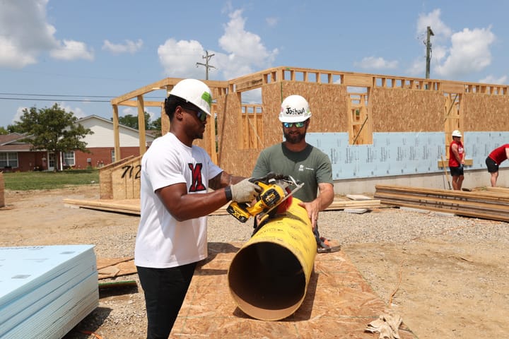 Dylan Downing stands on a construction site wearing a hard hat and using a saw to cut a large pipe