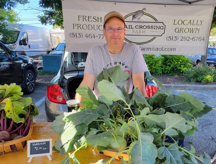 Greg Hamm stands behind a table with kohlrabi in a basket. The kohlrabi are round and green with leaves shooting out of 