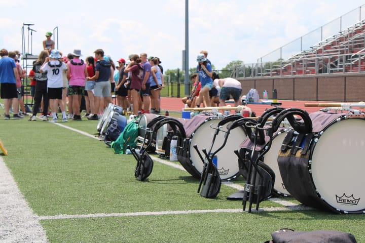 A set of bass drums is lined up on turf. A group of students stands in the background