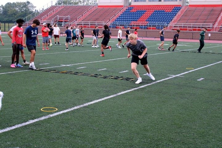 Football players run drills on the green turf in front of the blue and red stands. Photo by Taylor Stumbau