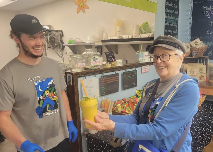 Jake Tyree wears a "Keep it Local: MOON Co-op" shirt. Next to him, Mitzi Ganelin holds up a yellow smoothie