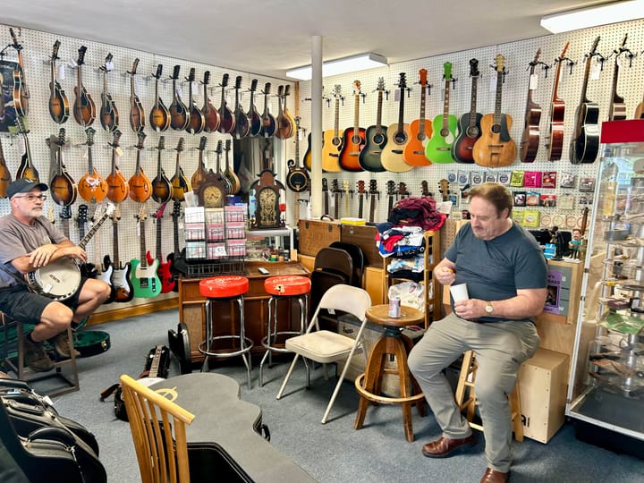 Two men sit in a room lined with different colored and types of guitars.