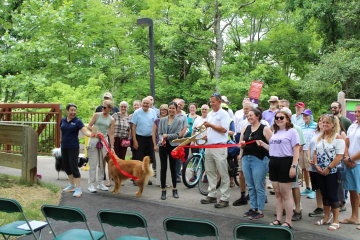 Dozens of community members stand on a trail as two people cut a large red ribbon