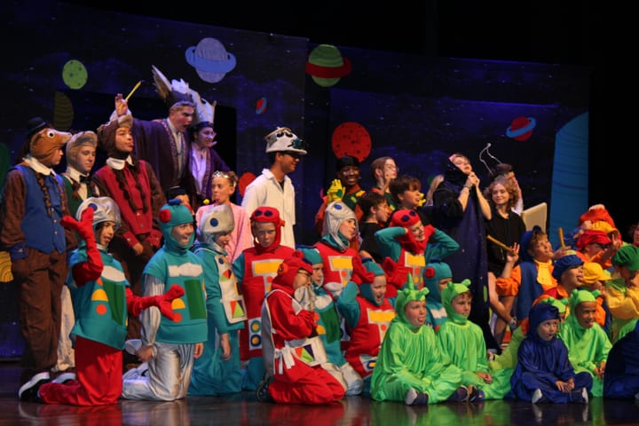 An ensemble of children in bright costumes pose for a cast photo on stage