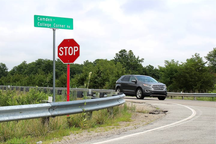 On the corner of two roads is a green road sign and a red stop sign with a black SUV driving by.