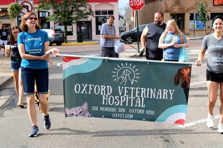 A group of men and women walk in a parade while holding a green flag with a dog and cat on it.