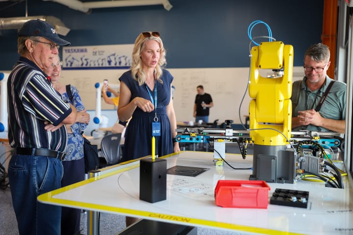 Several people look at a yellow robotic arm on a table