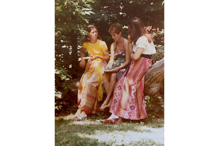 Three women sit on a log with plates and glasses in an old photo