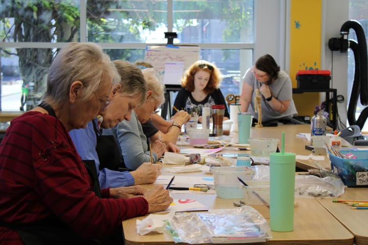 Several women paint at a cluttered table in a brightly lit room