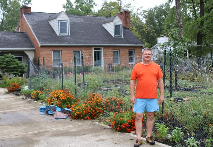 Jon Ralinovsky stands on his driveway in front of a fenced portion of his yard with many plants