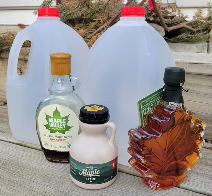 Two gallon jugs of water behind three brands of maple syrup