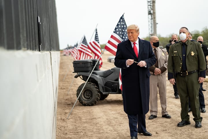 Donald Trump stands next to a border wall with American flags behind him