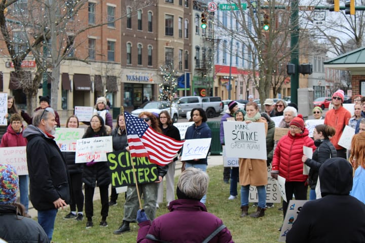 A woman holds an American flag in front of a group of protestors with science-related signs